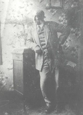 Studio photograph. An older man dressed in a suit stands leaning with his elbow against a cabinet. In the background are patterns of flowers.
