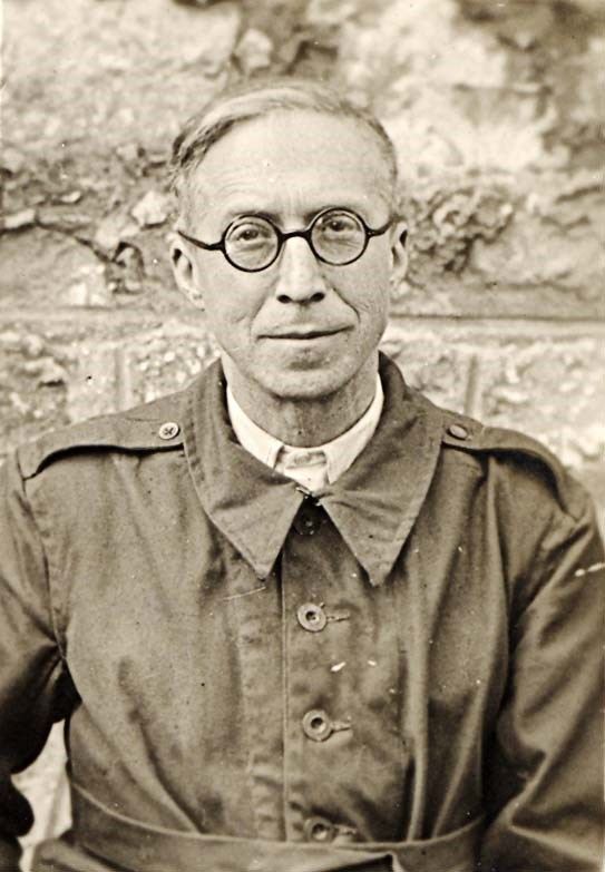 Black and white portrait of man in a coat, with round glasses, seated against a textured wall background.