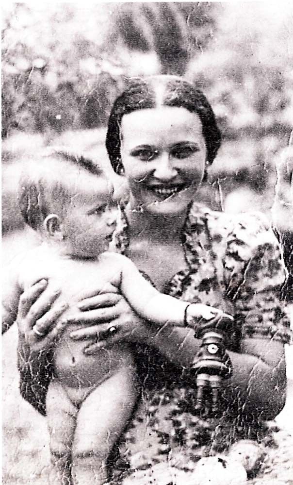 A vintage black-and-white photo showing a woman holding a small baby and a young child standing next to them, all smiling. The setting appears informal and possibly outdoors.