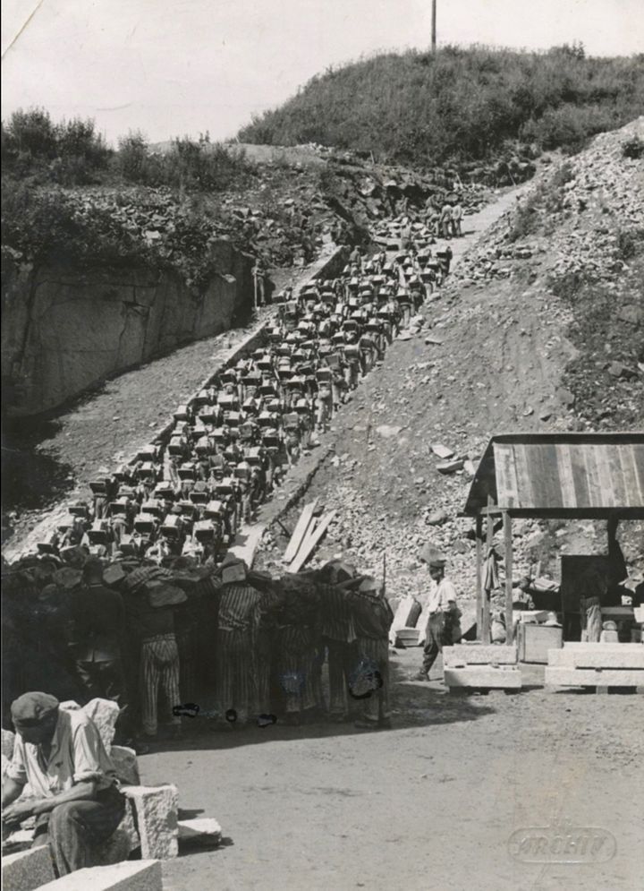 Historical black and white image of a large group of prisoners from Mauthausen concentration camp descending a narrow, rocky path at a significant slope. The individuals are carrying stones in baskets and containers on their backs. The surrounding environment is rugged with sparse vegetation, pointing to a hilly or mountainous region.