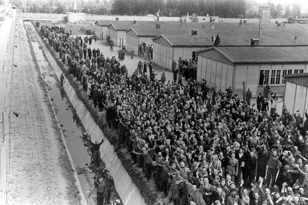 Crowds of prisoners of Dachau standing in front of the line of barracks. The fence of the camp is visible on the left.
