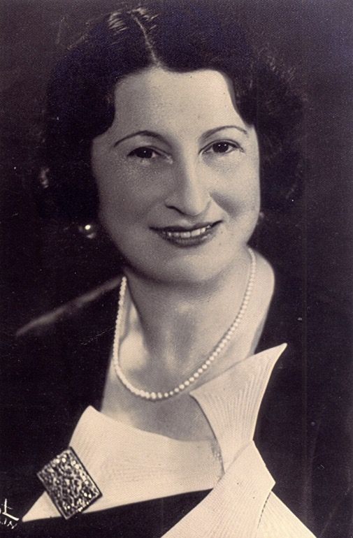 A portrait photo of a smiling woman with neatly styled long hair shaped in waves. She is wearing pearl beads and has some rectangular brooch at her blouse.