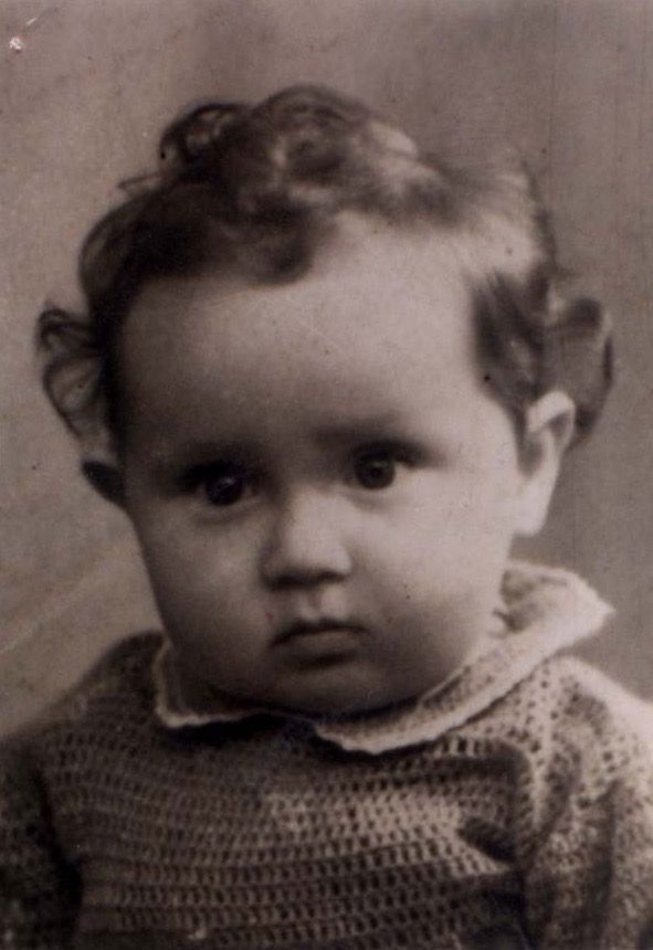Old black and white photograph of a young child with curly hair, wearing a knitted top. The child is looking directly at the camera with a serious expression.