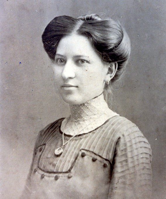 ortrait photo of a woman. Her hair is pinned up in buns on both sides of her head. She has a blouse with many embellishments, buttons and a voile fabric around her neck. She has a pendant around her neck.