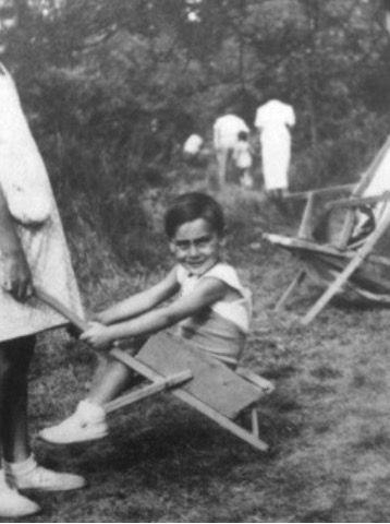 A boy sitting in a wheelbarrow being pushed by a slightly older girl. In the background is an empty recliner, a forest, and a group of people walking between the trees.