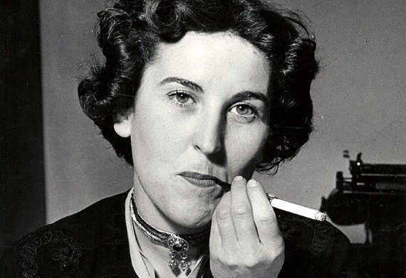 lack and white portrait of Charlotte Delbo, seated with a typewriter on a table behind her, holding a cigarette to her lips.