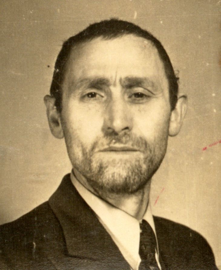 Portrait of an individual with a beard and mustache, wearing a dark suit and tie, set against a plain background.