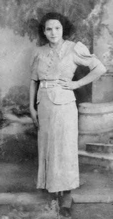 A black and white vintage photograph of a woman standing with one hand resting on the hip and the other on a pedestal.
