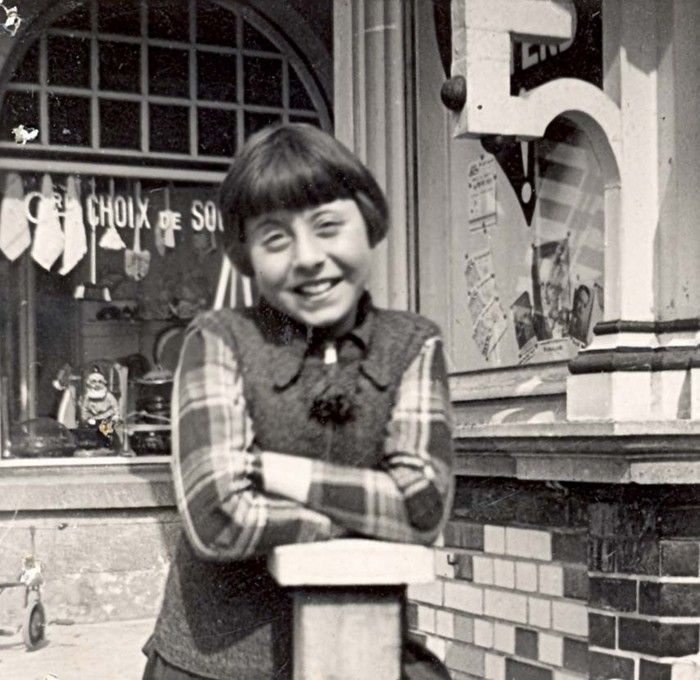 A girl smiling broadly, leaning on a railing in front of a store window with French text, "Choix de Souvenirs."