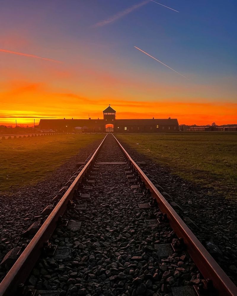 The iconic railway tracks leading into the Auschwitz-Birkenau concentration camp, framed under a vibrant orange and blue sunset sky. The tracks converge at the main entrance gate with the guard tower silhouetted against the fiery hues of the setting sun.