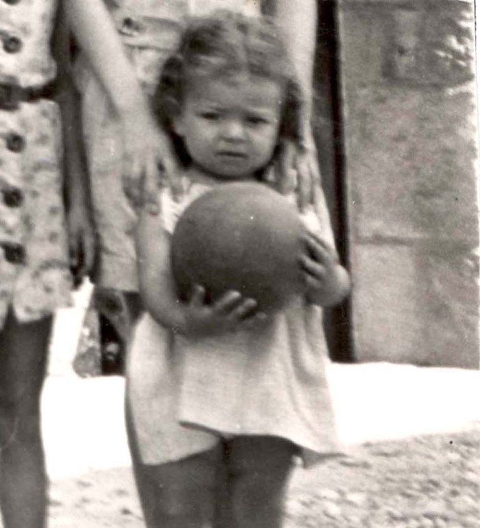 A vintage black and white photo showing a young girl standing and holding a large ball. The child has curly hair and is wearing a light-colored outfit. Only part of an adult, standing next to the child and dressed in a light-colored outfit, is visible on the left side of the frame. The background shows a nondescript, blurry outdoor setting.