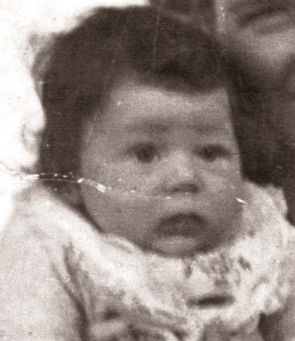 Close-up of a baby with wide eyes and a slightly open mouth, wearing a textured outfit. The background is blurry, partially showing another person's face. The image is monochrome and has visible signs of aging and wear.