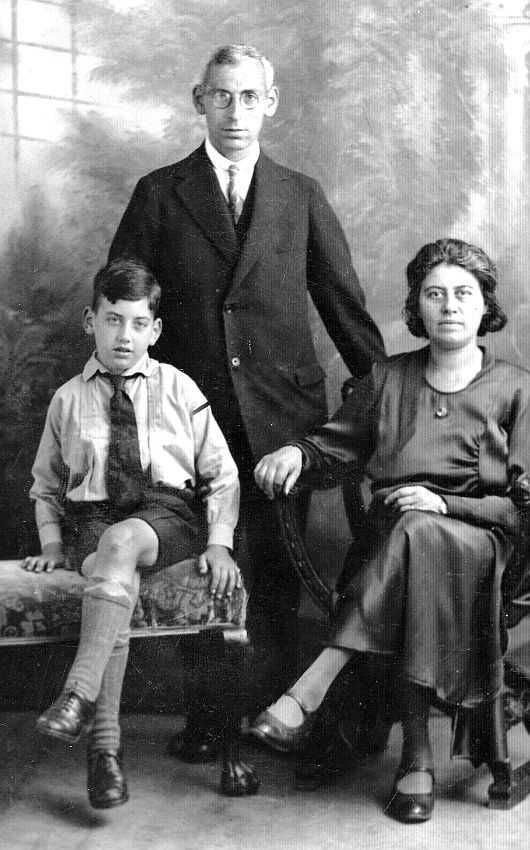Vintage black and white photo of three family members posing for a portrait. One child seated on a bench to the left, a man standing in the center, and a woman seated on the right. They are all dressed in formal early 20th-century attire.