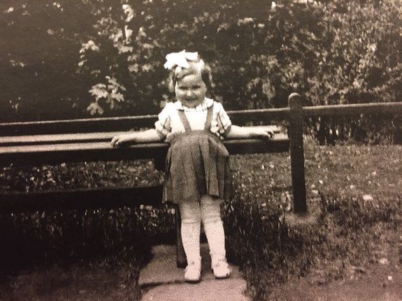A vintage black and white photo of a young girl standing in front of a wooden fence with lush foliage in the background. She is wearing a dress and a bow in their hair, and is smiling at the camera.