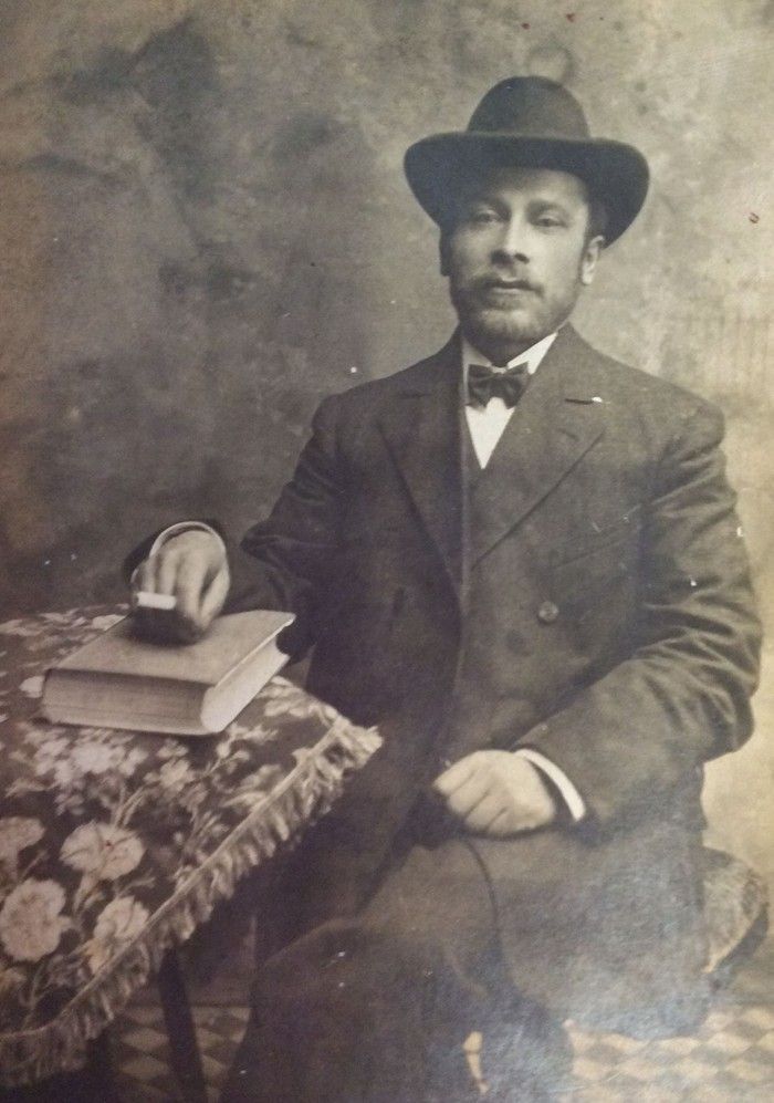 A vintage photograph of a person in formal attire with a bowtie, holding a book and standing next to a small table with a patterned tablecloth. The person is wearing a hat and has a well-groomed mustache. The background is plain.