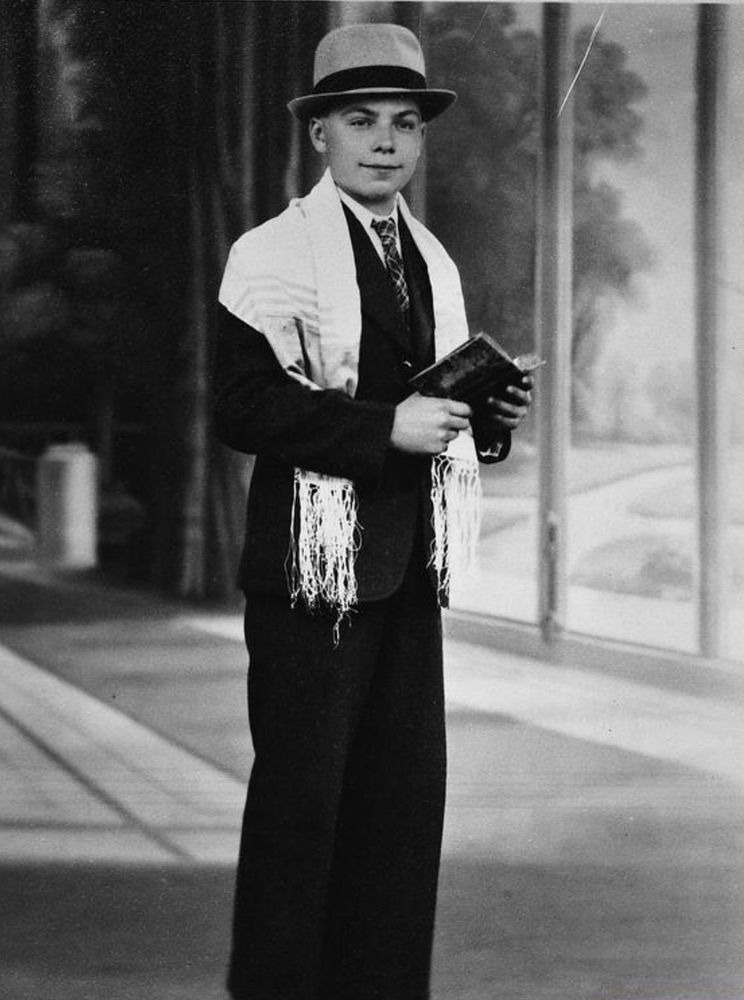 A young person from the early 20th century dressed in a formal outfit, holding a book and wearing a jacket, dark vest, bow tie, and a straw boater hat. He has a Jewish prayer shawl on his shoulders.