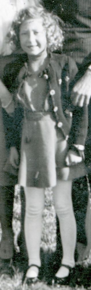Old black-and-white photo of a young girl standing outside. The child is dressed in a coat with decorative buttons and a skirt. She has curly hair. The background is mostly indistinct, suggesting a group setting. The focus is on the child's smiling expression.