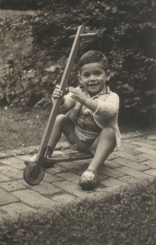 A vintage black and white photo of a young childboy smiling and sitting on a brick pathway, on a scooter. The background features a garden with visible foliage.