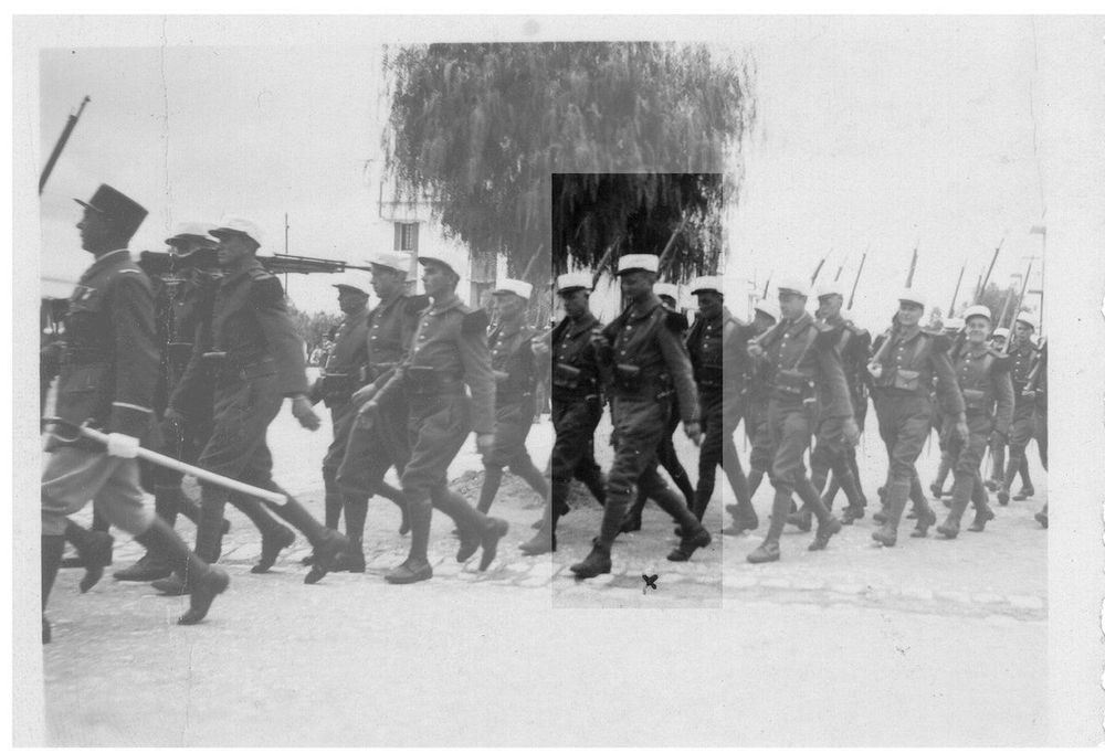 A historical black-and-white photograph of a group of uniformed soldiers marching in formation. One soldier at the front carries a flag, while others carry rifles. The setting appears to be an open area with a sparse tree in the background.