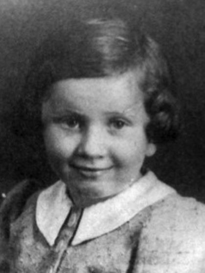 Portrait photograph of a young girl against a solid background. You can see her face and her eyelashes. She is wearing a buttoned-up neck blouse with a collar. She is smiling slightly. She has medium-length, slightly wavy hair.