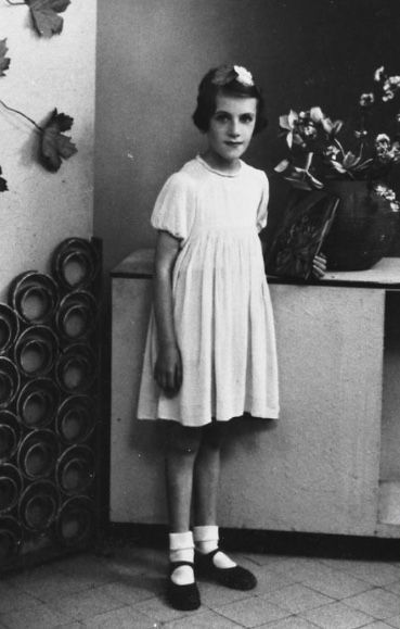 A girl standing in a room, wearing a white dress, white socks, and black shoes, with a flower in the hair.