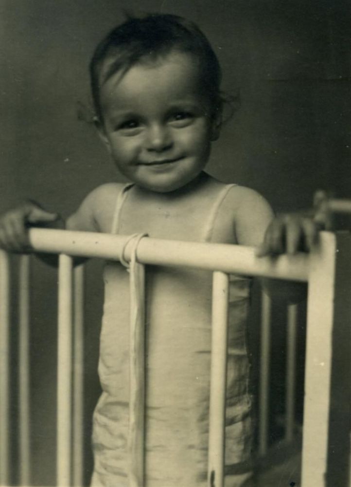 A girl standing in a crib - leaning against the rungs of the crib wall.