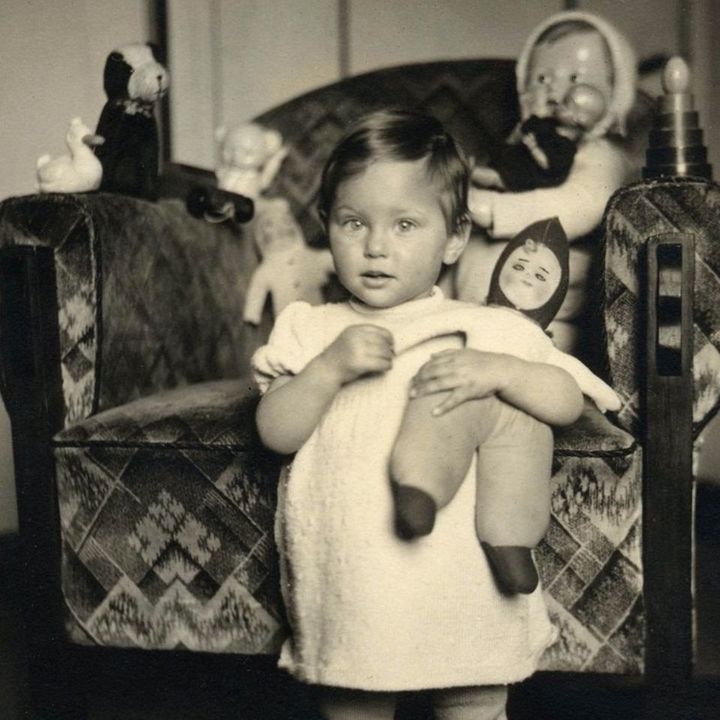 A vintage black and white photo of a young girl wearing a dress, standing in front of an armchair adorned with a patterned fabric. The child is surrounded by various toys, including dolls.
