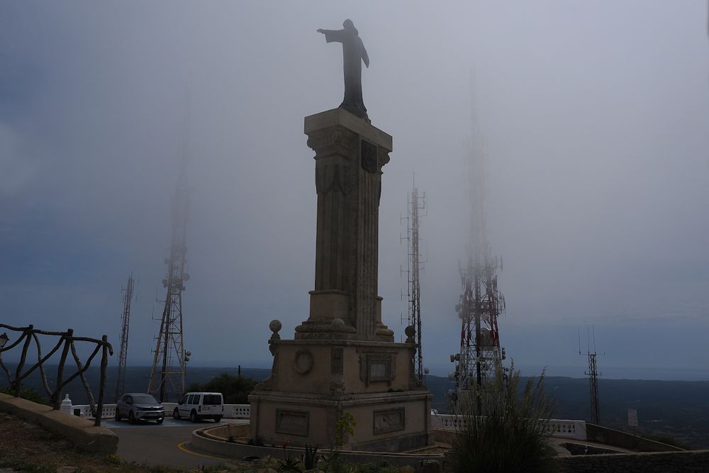 Photo: a statue of Jesus Christ on a very tall pedestal, seen from behind & below, with its arms spread wide, surrounded by radio antennas and cellphone towers, in a small parking area before the Sanctuary of the Virgen del Toro (not shown) that stands atop El Toro, the highest point on the Balearic Island of Menorca. It is a mostly cloudy day and fog obscures the communication towers. A car and a van are parked near the base.

Mirador Cim del Toro, Menorca
Balearic Islands, Spain
April 2023
OM-1: ƒ/11, 1/1250, 14mm, ISO200