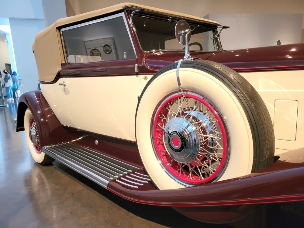 Photo: In a museum gallery, a side view from the front right corner of the passenger side of a 1931 Packard Model 840 Dietrich Convertible Victoria. The fenders are a dark red swooping over the tires, which are creamy white white walls to match the door and side panels. The running board is the same dark red, except for the step portion beneath the door, which is chrome. A spare wheel is mounted just forward of the passenger door, along the long hood. The hub caps are chrome, of course, with Packard branding at the hub and a complex chrome spoke system connecting it to the rim and tire. The rim is a brighter red, matching the pin striping on the fenders.
Nelson Atkins Museum of Art
Kansas City, Missouri
July 2022