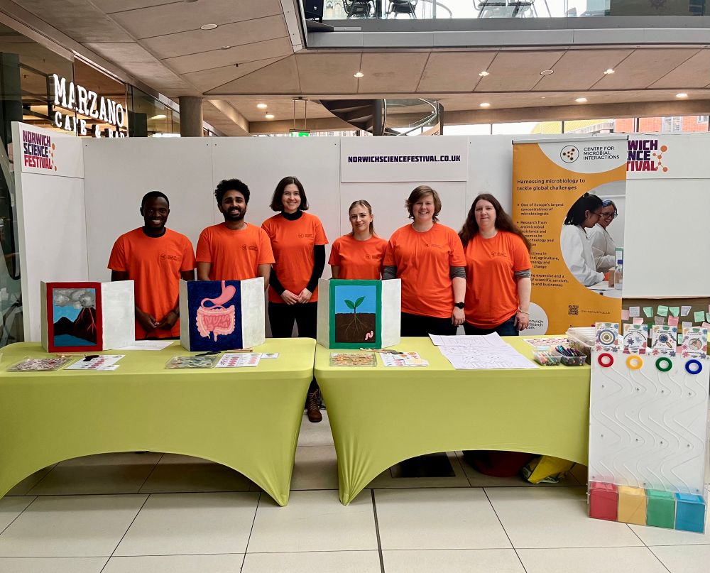 A group of six volunteers at Norwich Science Festival. They are wearing bright orange t-shirts and standing behind two tables displaying microbiology outreach activities for the public