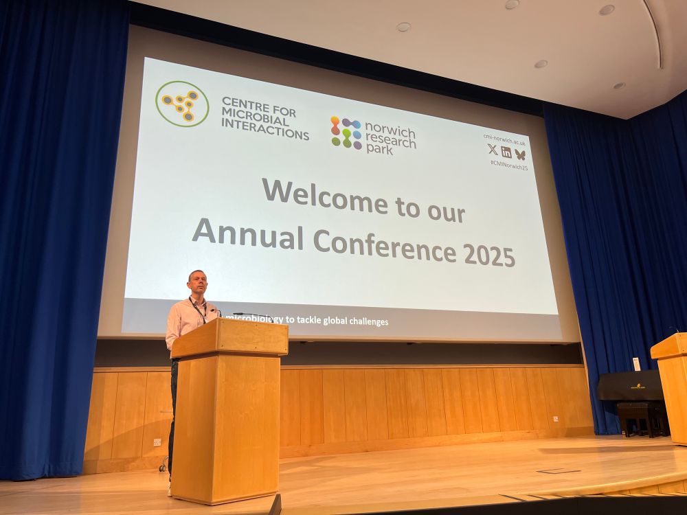 Mark Webber standing behind a podium on a conference centre stage. The large screen behind him says ‘Welcome to our Annual Conference 2025’.