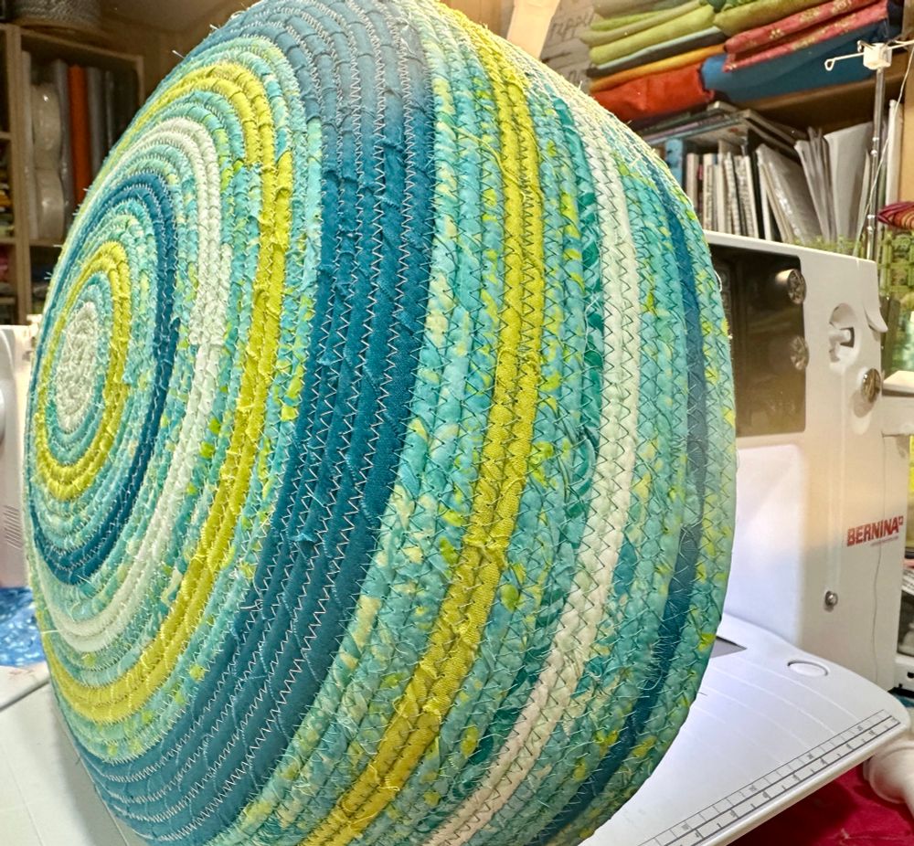A large rope basket on a sewing machine. The spirals of rope are wrapped in bright colors: aqua, teal, and acid yellow. Its base is 14” and the sides are about 4”, but will become much deeper (until I run out of fabric). 