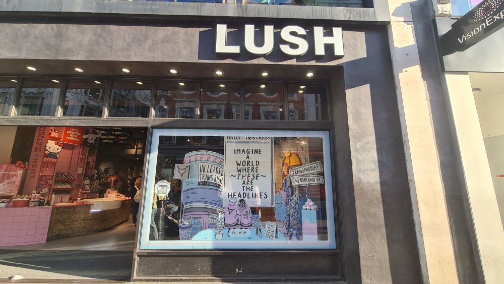 Picture of the storefront of a Lush store with a display focused on a newspaper saying Imagine a world where these are rhe headline, a picture of two peolle with their backs to the viewer, with a trans flag hat ans logo, and news headlines surrounding them such as UK leads on trans rights, unrealistic waiting times trashed, GPs on board with gender affirming care, trans youth living their truth, basic trans healthcare in the bag, the UK a safe haven for lgbtq+ asylumn seekers and transphobia, we don't know her.