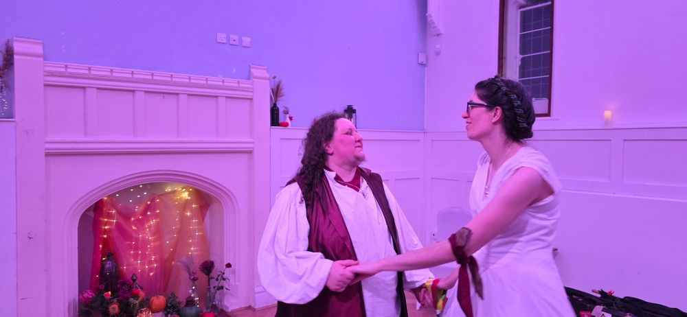 Charley in a red waistcoat and white shirt with long curled hair dancing with Jude in a long white dress, gazing at each other, in front of a fireplace full of fairly lights and flowers