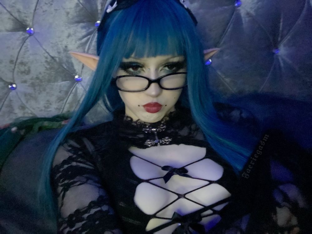 Avilone pouts at the camera for a selfie. They have dark blue long hair, elf ears, big dark eyeliner, dark red lipstick and black piercings. They have on a black mesh dress which reveals the middle of their chest with bows down the front and long lace sleeves.