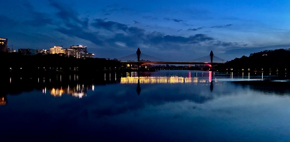 A pastel-hued cityscape with a glowing bridge reflected in calm lake waters under a soft twilight sky