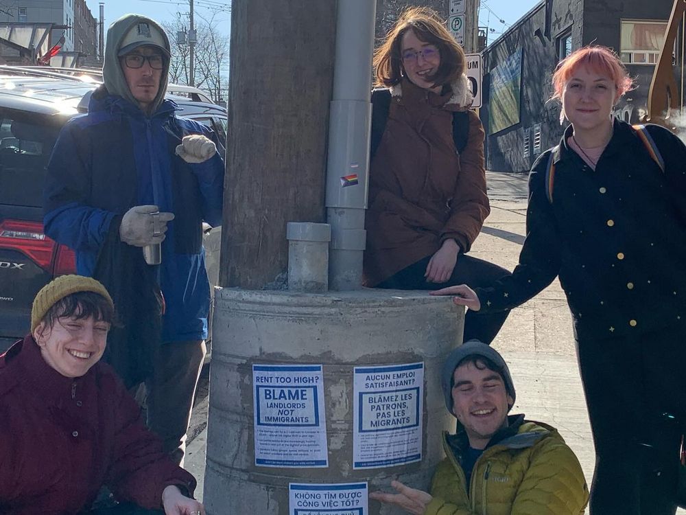 Five people on a street in Halifax standing around three wheat-pastered DON'T BLAME IMMIGRANTS flyers, smiling. 
