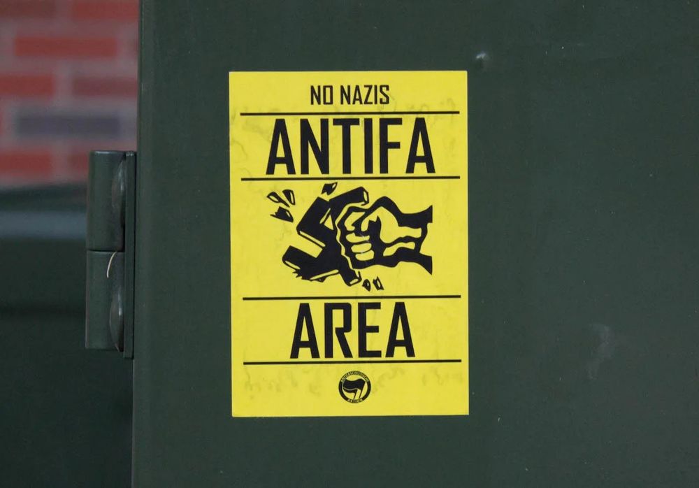 Yellow sticker on a utility box, showing a fist smashing a swastika, captioned NO NAZIS - ANTIFA AREA 