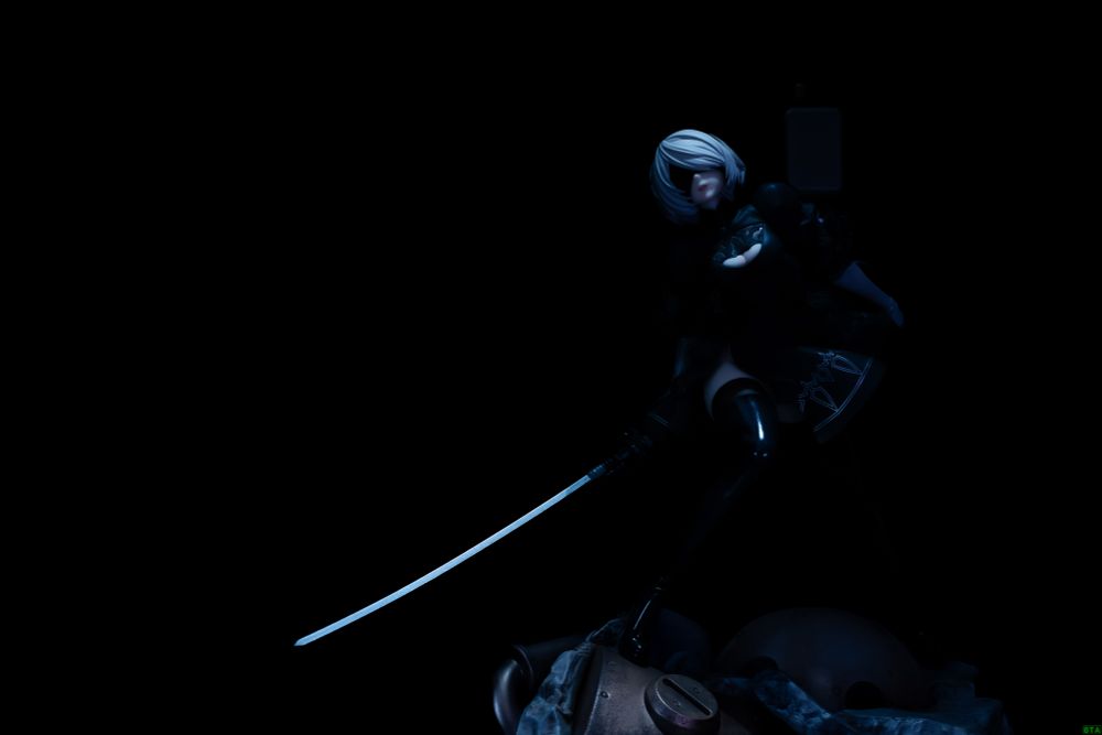 2B emerges from the darkness. this photo captures both the pitch black areas and the well-lit places that are intended to be shown off. The way each section is lit up is also excellent, such as the gleam of the sword, the soft shimmer of her hair, the bright sparkling skin of her chest and thighs showing from her boots, and a faint light filtering onto the base. Even though most of her body is shrouded in darkness, this photo really stirs your imagination.
