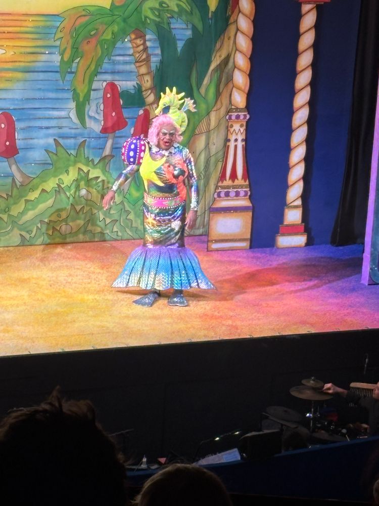Performer dressed in a colorful, glittering costume on a theater stage with a tropical-themed backdrop, featuring palm trees, mushrooms, and columns. The performer wears a shimmering dress, pink wig, and elaborate accessories, engaging with the audience during a lively show.