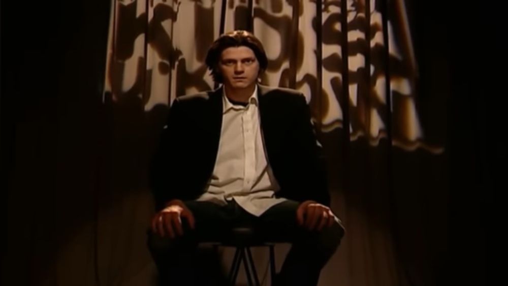 The late Trevor Moore sitting in a chair and looking at the camera with a stern expression, as if demonstrating a severe law. 