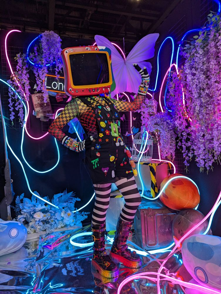 A 'TV Head' cosplayer in a Kidcore-themed outfit poses in a neon vaporwave photobooth.