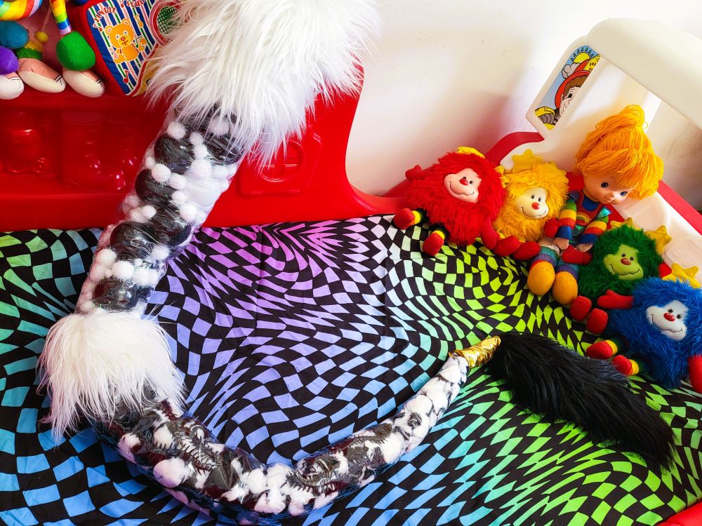 Photo of a long serpentine fursuit tail with a black & white colour scheme, featuring transparent sections filled with various objects such as ball-pit balls, pom-poms and plastic bugs.