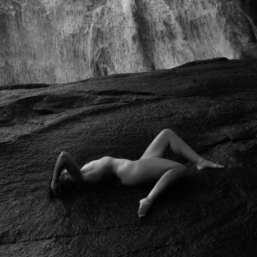 A nude woman reclines on black rock at the base of a waterfall, her legs and arms making triangular patterns. The image is in black and white.