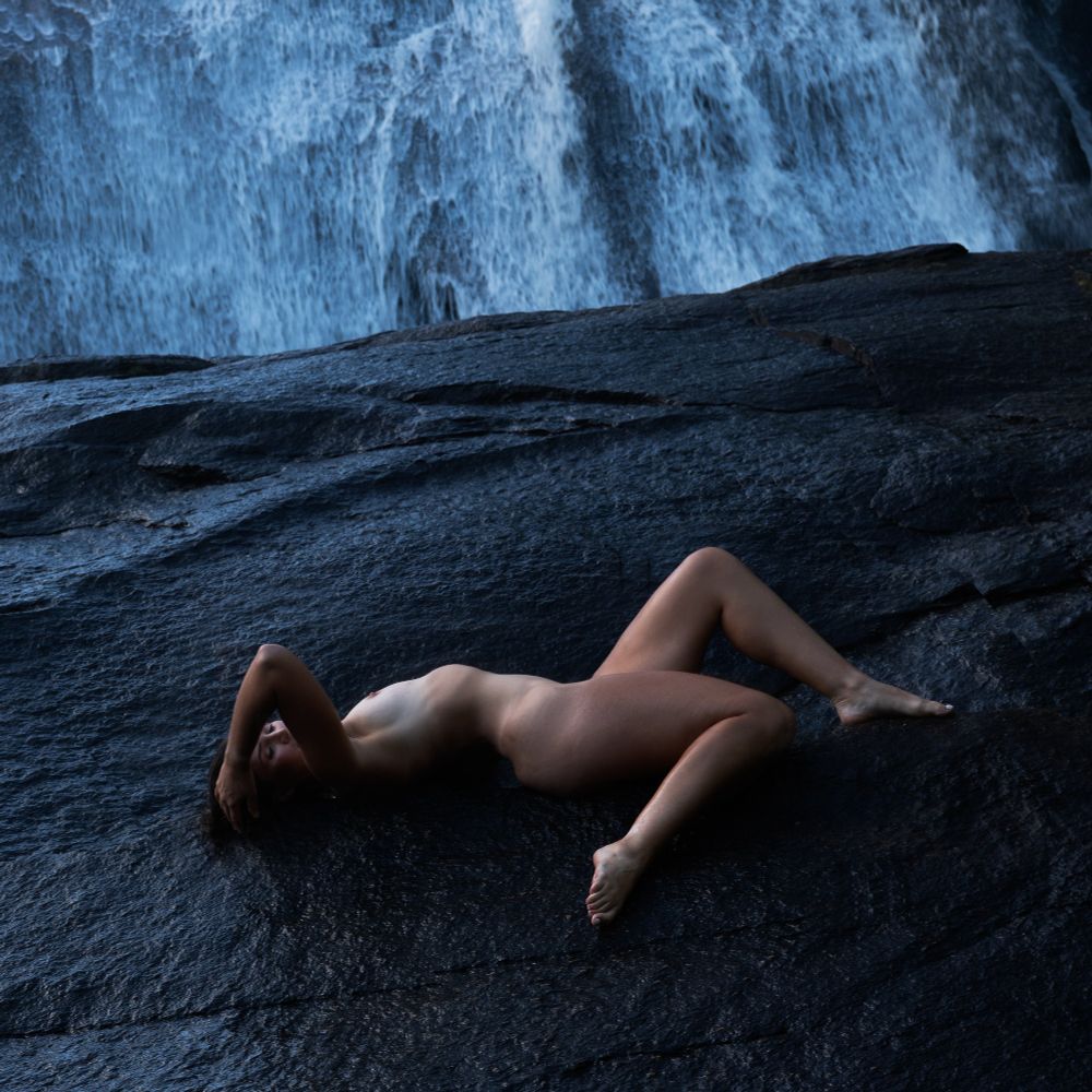 A nude woman reclines on black rock at the base of a waterfall, her legs and arms making triangular patterns. The color image has a cool blue background that contrasts her warmer skin tone.