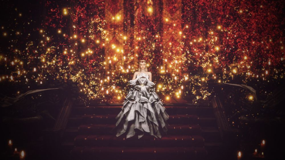 Aristen stands in the middle of the stairs in an elegant white dress, her hands on her chest. She is against a yellow and red bokeh background. She has a wreath of red roses on her head.