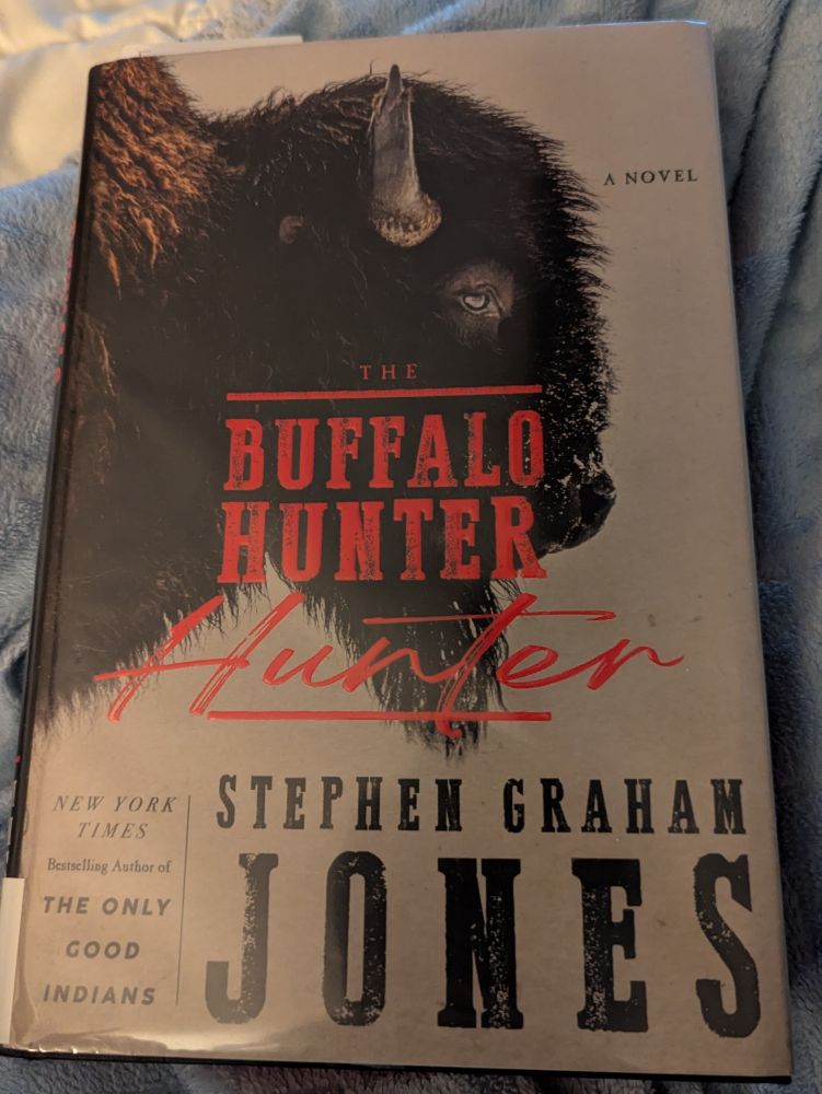A physical copy of "The Buffalo Hunter Hunter" by Stephen Graham Jones lying on top of a light blue blanket