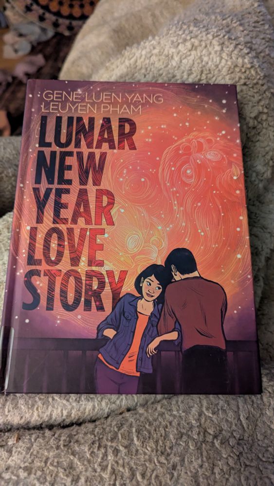 A copy of "Lunar New Year Love Story" by Gene Luen Yang and Leuyen Pham sitting on a beige fuzzy blanket.