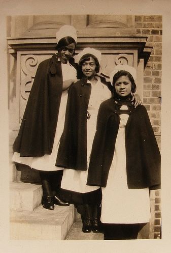 three Black women, assumed to be nurses, standing on three steps. they are all wearing white nurse uniforms and a black coat over their shoulders that end near their thighs. they are all wearing black stockinfs and black shoes. they are also wearing white nurse hats. they are smiling and have hands on each others shoulders