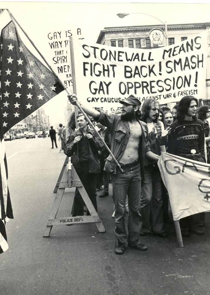 Photo sépia de Léonard Finck représentant un groupe de manifestants gays états-uniens lors d'une manifestation en 1969 suite aux émeutes de Stonewall. La personne au premier plan tient la banderole de tête et un drapeau des usa (hors champ). Derrière lui une banderole où on peut lire : "Stonewall signifie la riposte ! La lutte contre l'oppression des homosexuels ! Signé un groupe gay contre la guerre et le fascisme"